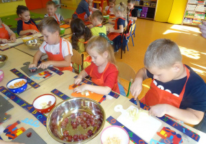 Troje dzieci siedzi przy stole nakrytym podkładkami z rozłożonymi deskami, w ręku trzymają noże, którymi kroją owoce. Na środku stołu stoją miski z owocami.